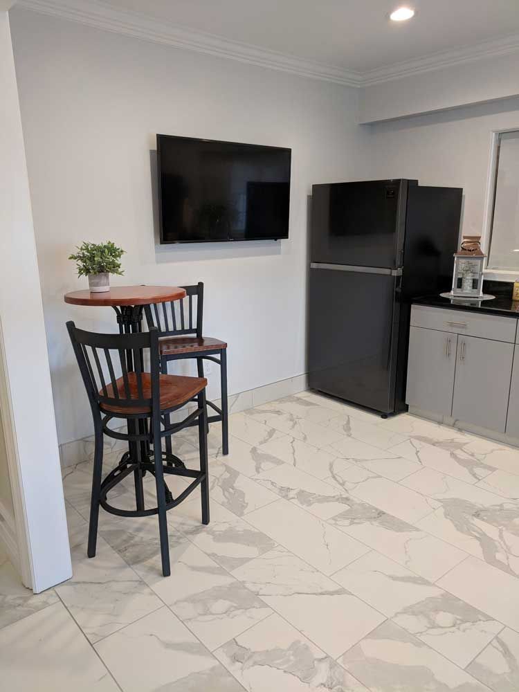 A small dining area with a round table, two black bar stools, a television, a black refrigerator, and marble tile floors.