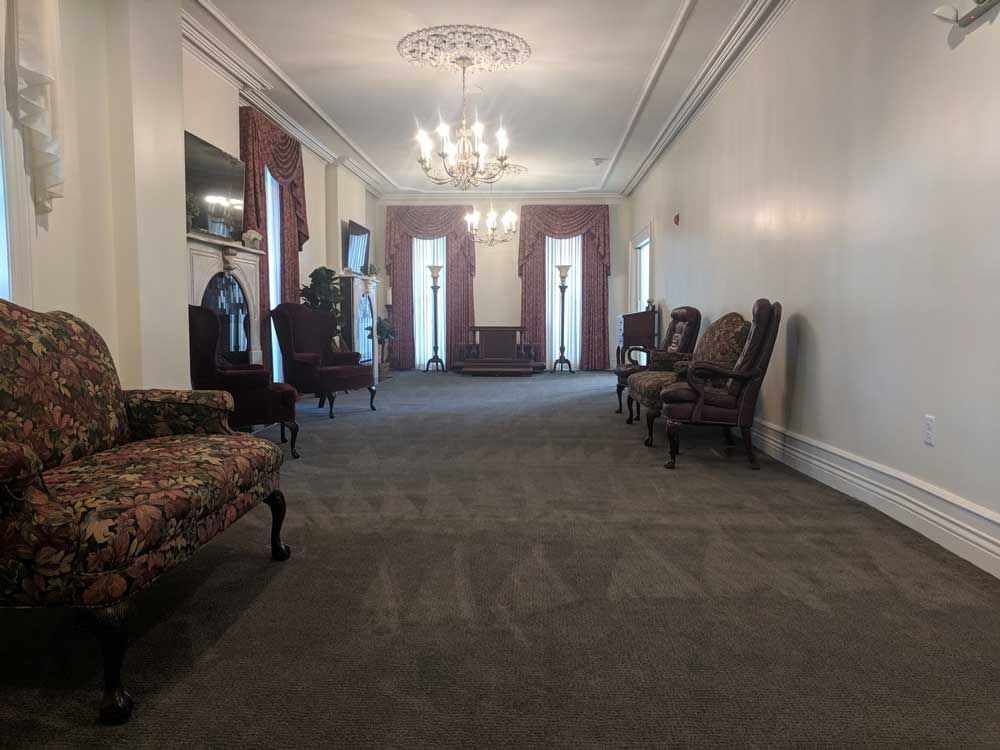 A formal, carpeted parlor room features several ornate floral armchairs, a fireplace, and crystal chandeliers.