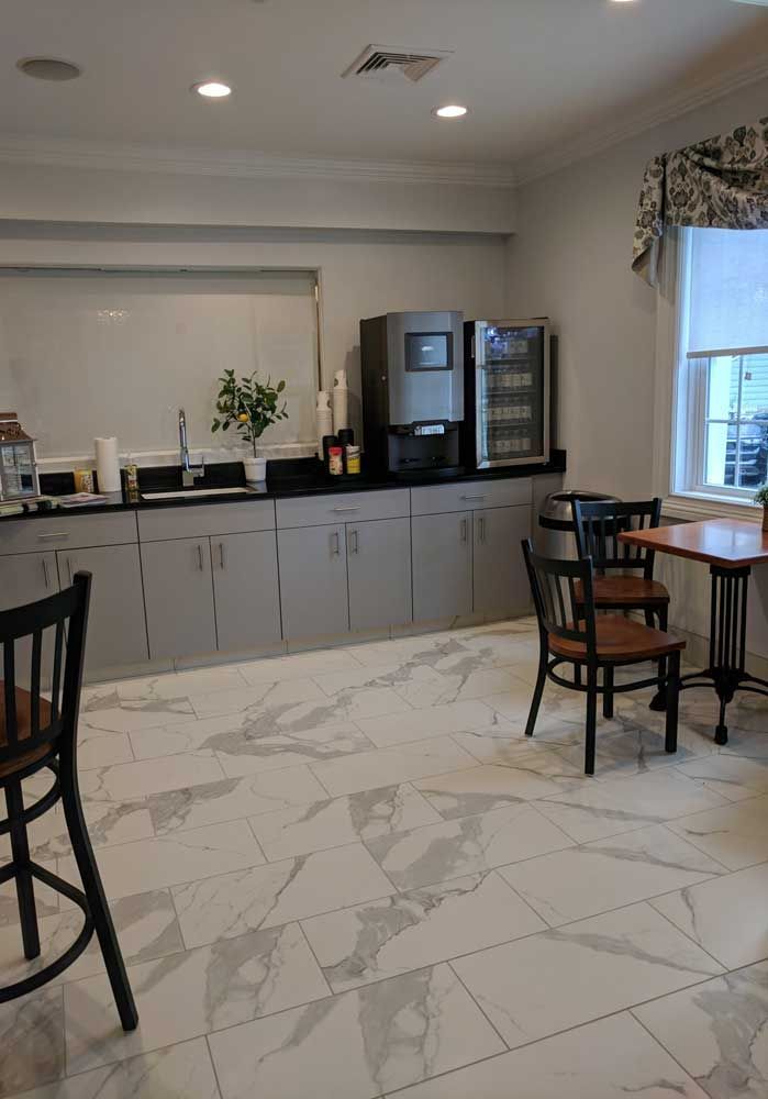 A refreshment station with a coffee machine, gray cabinets, white marble-patterned flooring, and a table with chairs.