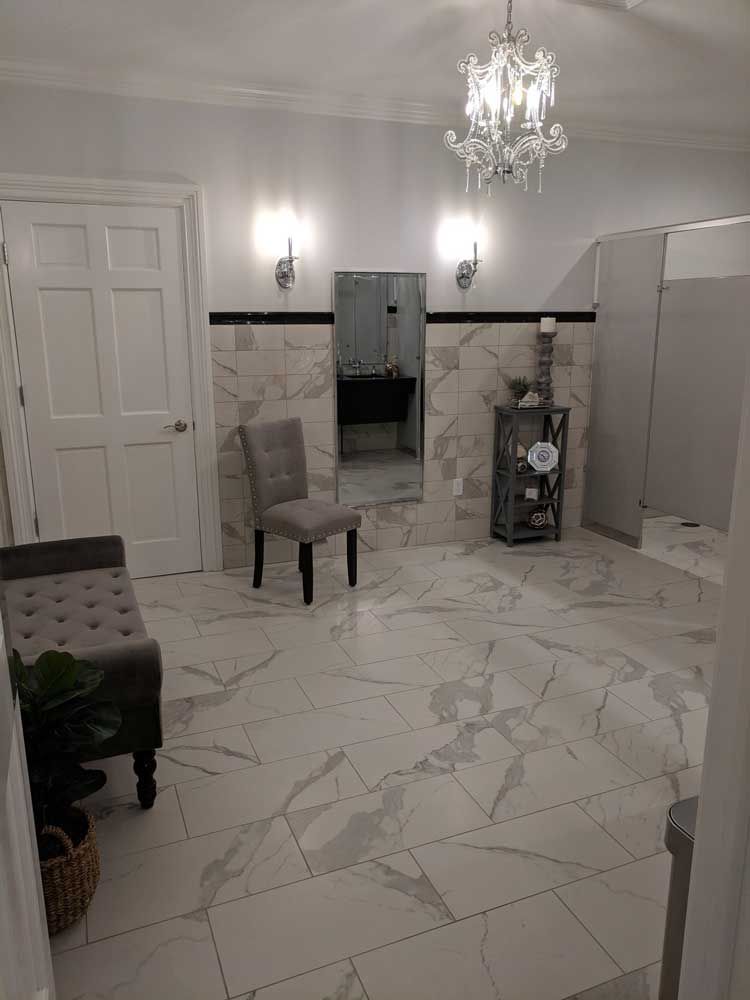 A brightly lit bathroom with white marble tile floors and walls, a crystal chandelier, a gray chair, and a white door.