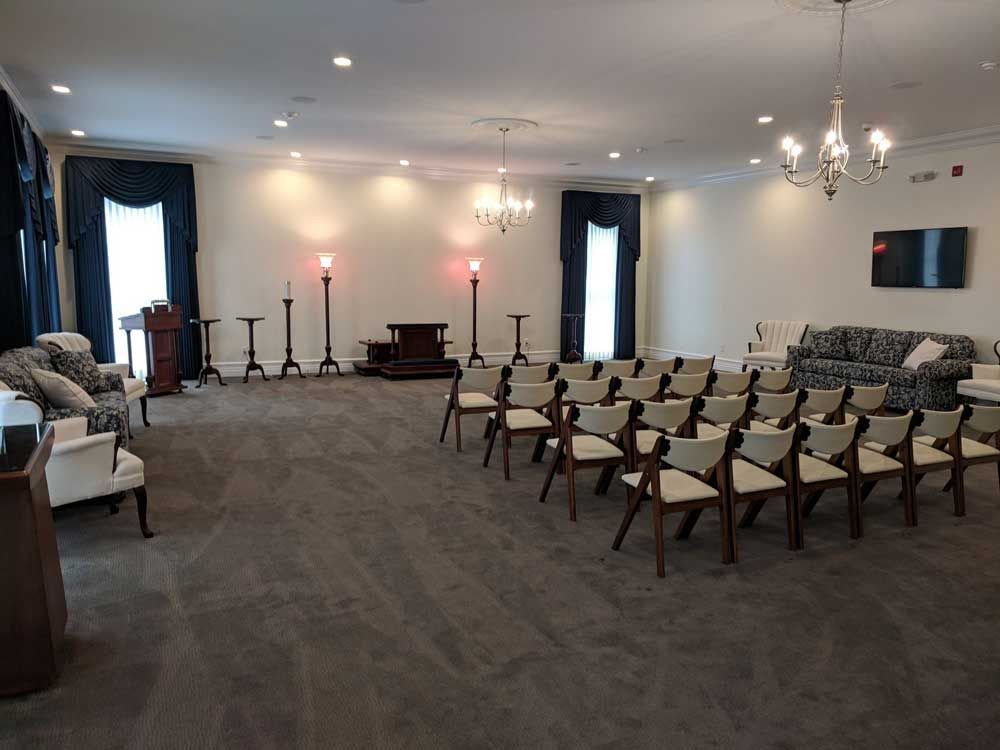 A brightly lit, neutral-toned room with chairs in rows, a sofa, dark drapes, chandeliers, and a small podium.