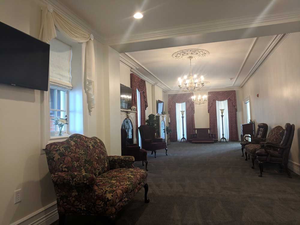 A spacious, formal room with floral furniture, elegant drapes, chandeliers, and dark carpeting in a neutral-toned setting.