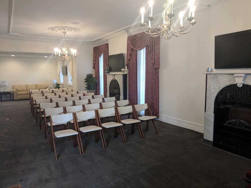A formal room arranged with rows of white chairs facing a fireplace with a mounted television and burgundy curtains.