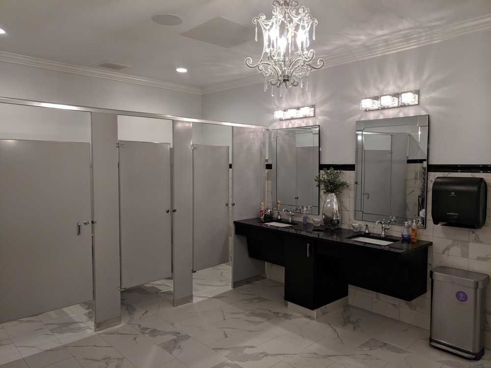 A modern restroom with a chandelier, two mirrors over a black vanity, gray stall doors, and a paper towel dispenser.