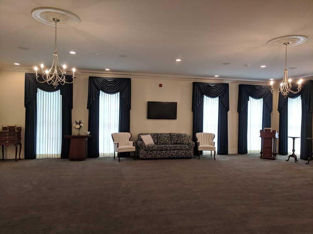 A formal room with neutral carpet, two chandeliers, a patterned sofa, two armchairs, and dark draped windows.