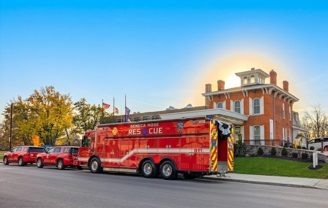 A large red rescue fire truck parked in front of a historic red brick house during a sunset.
