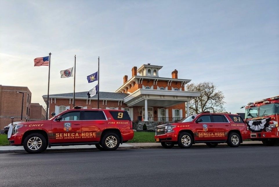 Red fire department vehicles parked in front of a historic brick building with flags at half-staff.