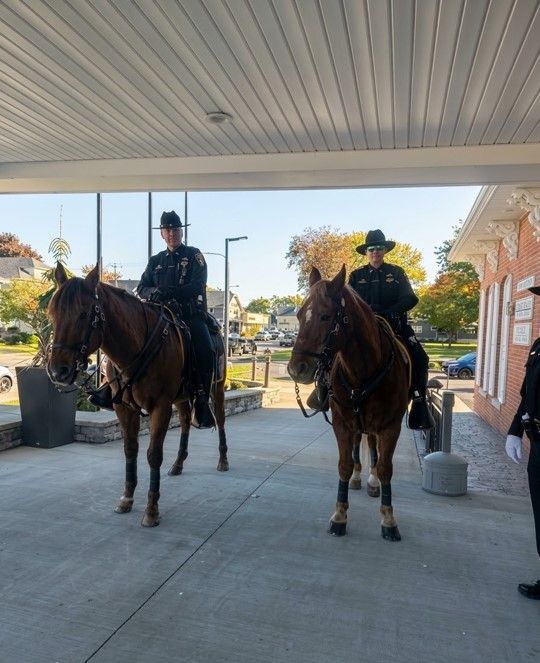Two mounted police officers in uniform and hats sit on brown horses under a building overhang on a sunny day.