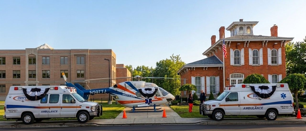 Two ambulances and a medical helicopter adorned with patriotic bunting parked in front of a red brick historic building.