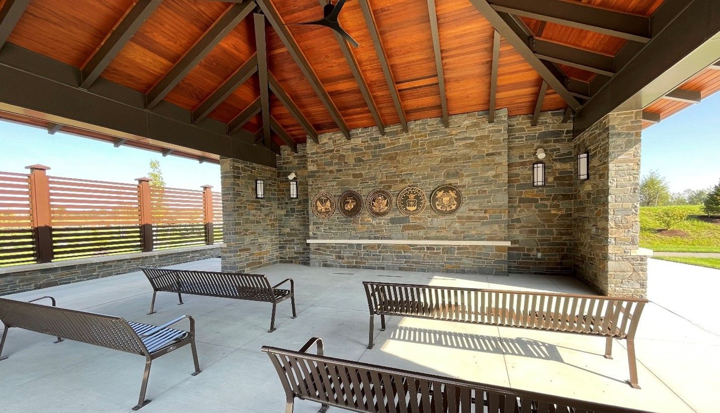 An open-air pavilion with stone walls, a wood-beam ceiling, and metal benches arranged on a concrete floor.