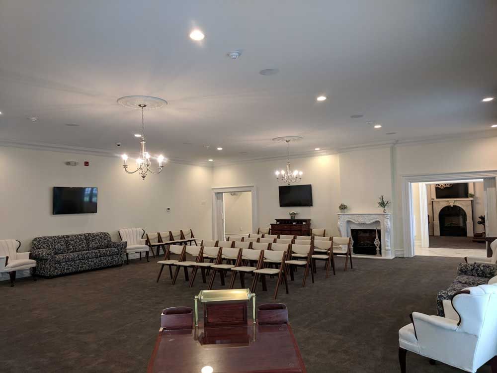 A spacious room set up for a gathering, featuring rows of chairs, a couch, armchairs, two fireplaces, and chandeliers.