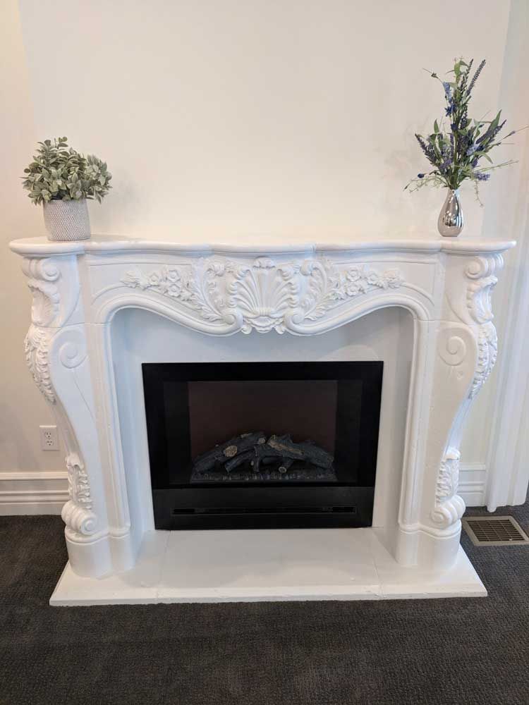 A white, ornate carved fireplace mantel with an electric insert, featuring small potted plants on the mantle shelf.