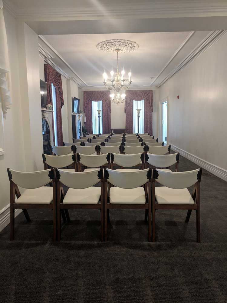 A room set up with rows of white chairs facing windows with draped curtains and a hanging chandelier.
