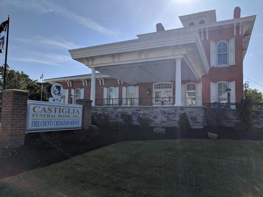Castiglia Funeral Home building, a brick structure with white columns and a large sign in the foreground, under blue sky.