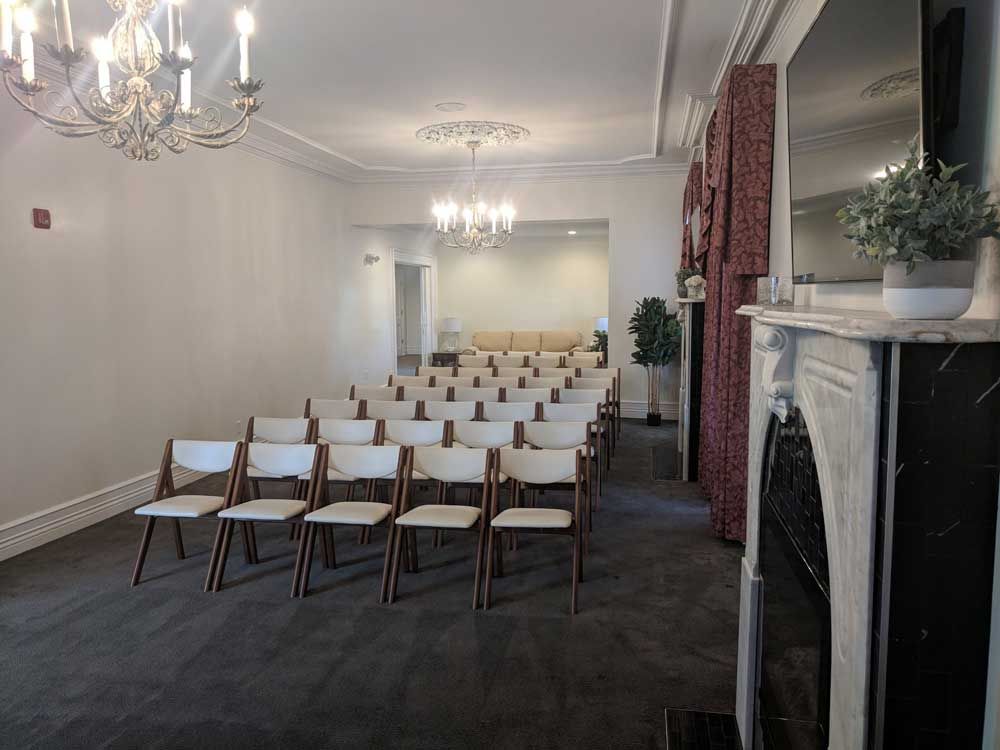 A dimly lit event space featuring rows of white chairs facing a distant couch, illuminated by two ornate chandeliers.