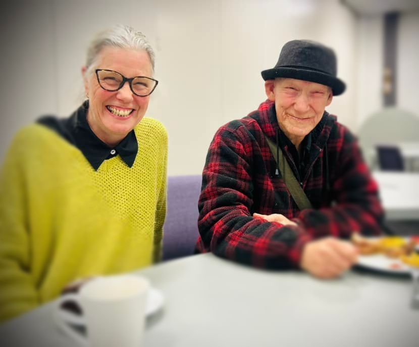 Woman in yellow sweater smiles at camera next to man in plaid shirt and black hat, at a table.