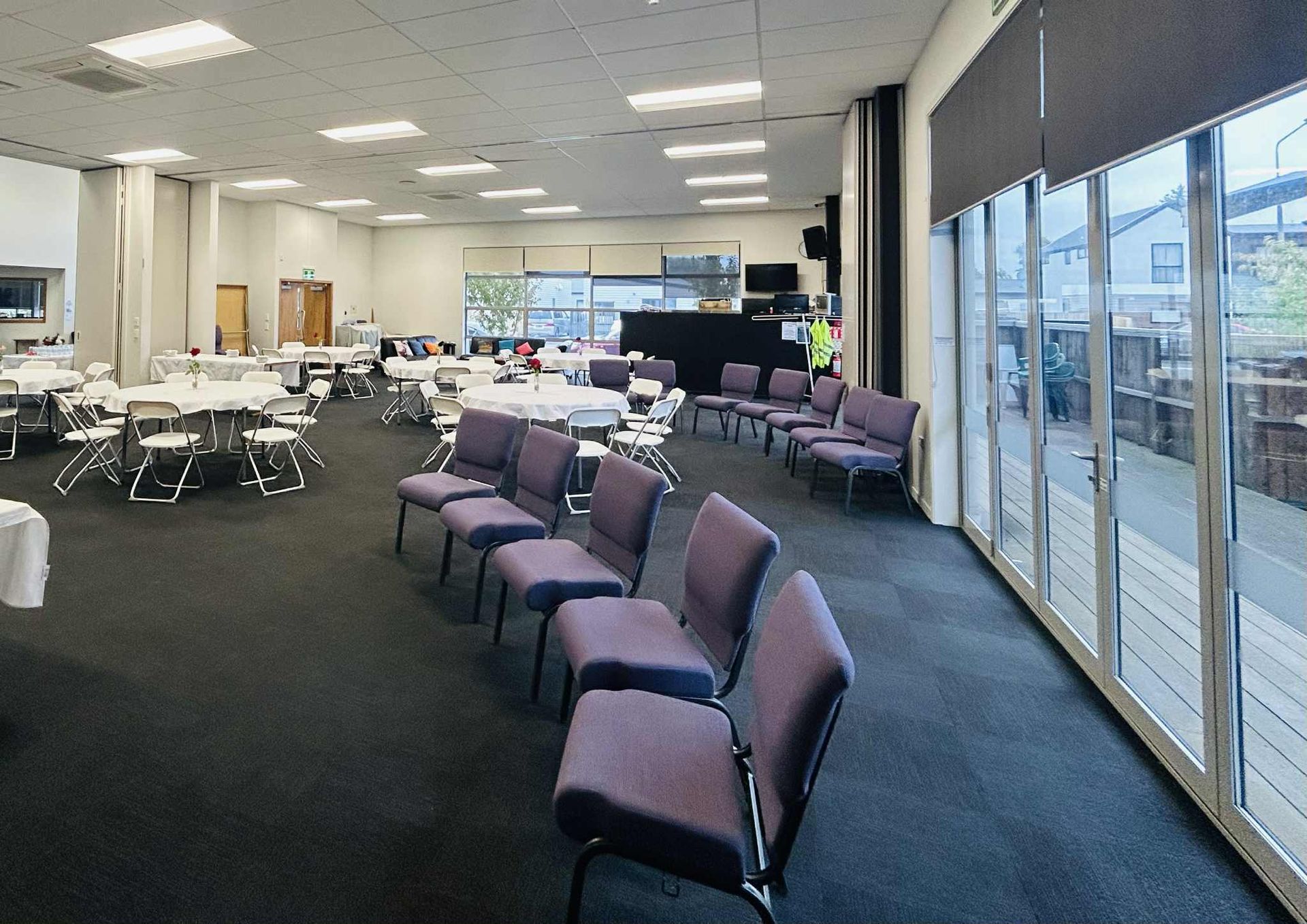 A large event space with tables and chairs, featuring rows of purple chairs and large windows.