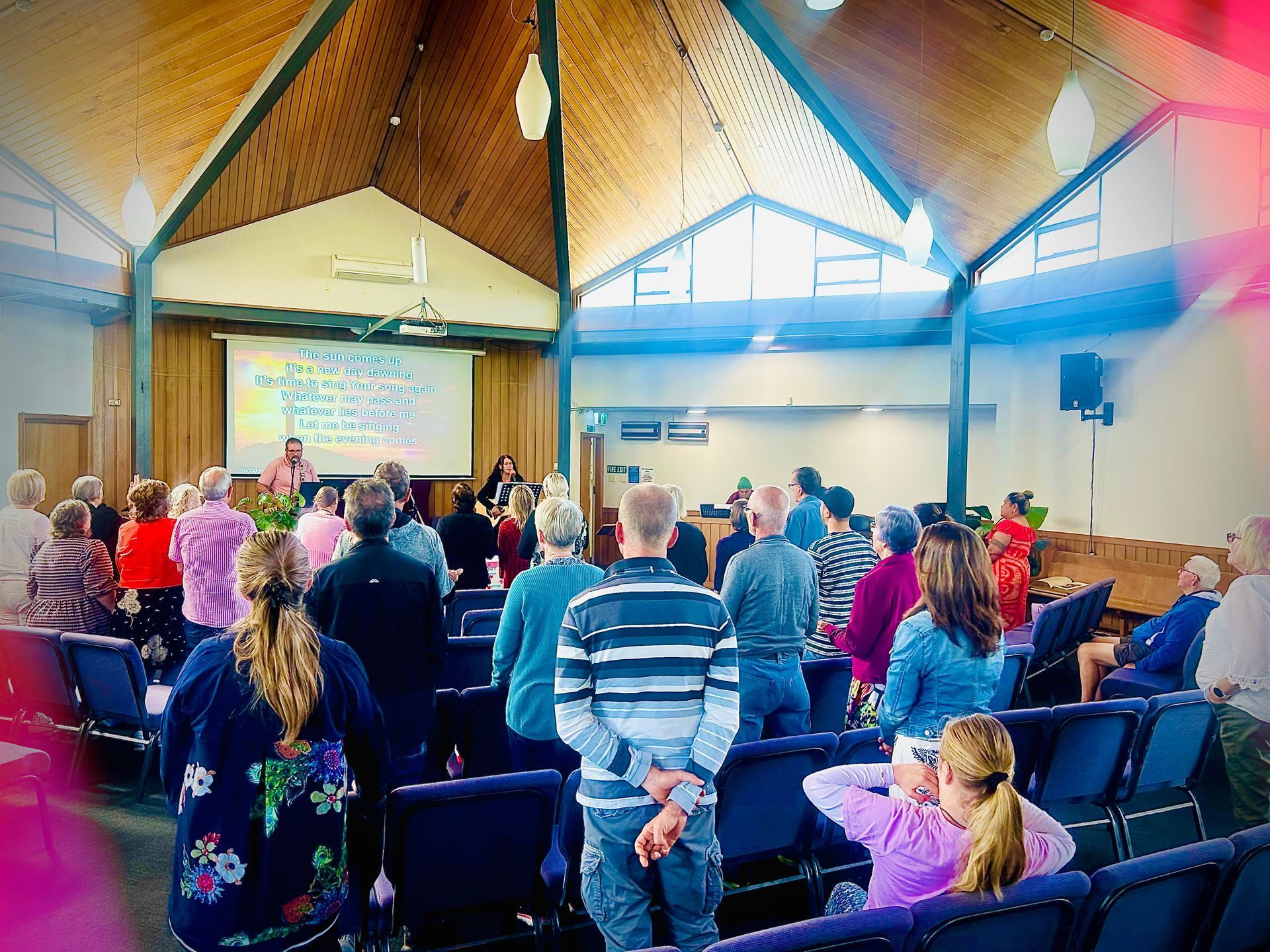 People gathered in a church for a service. Wooden ceiling, stage with a presentation, attendees singing.