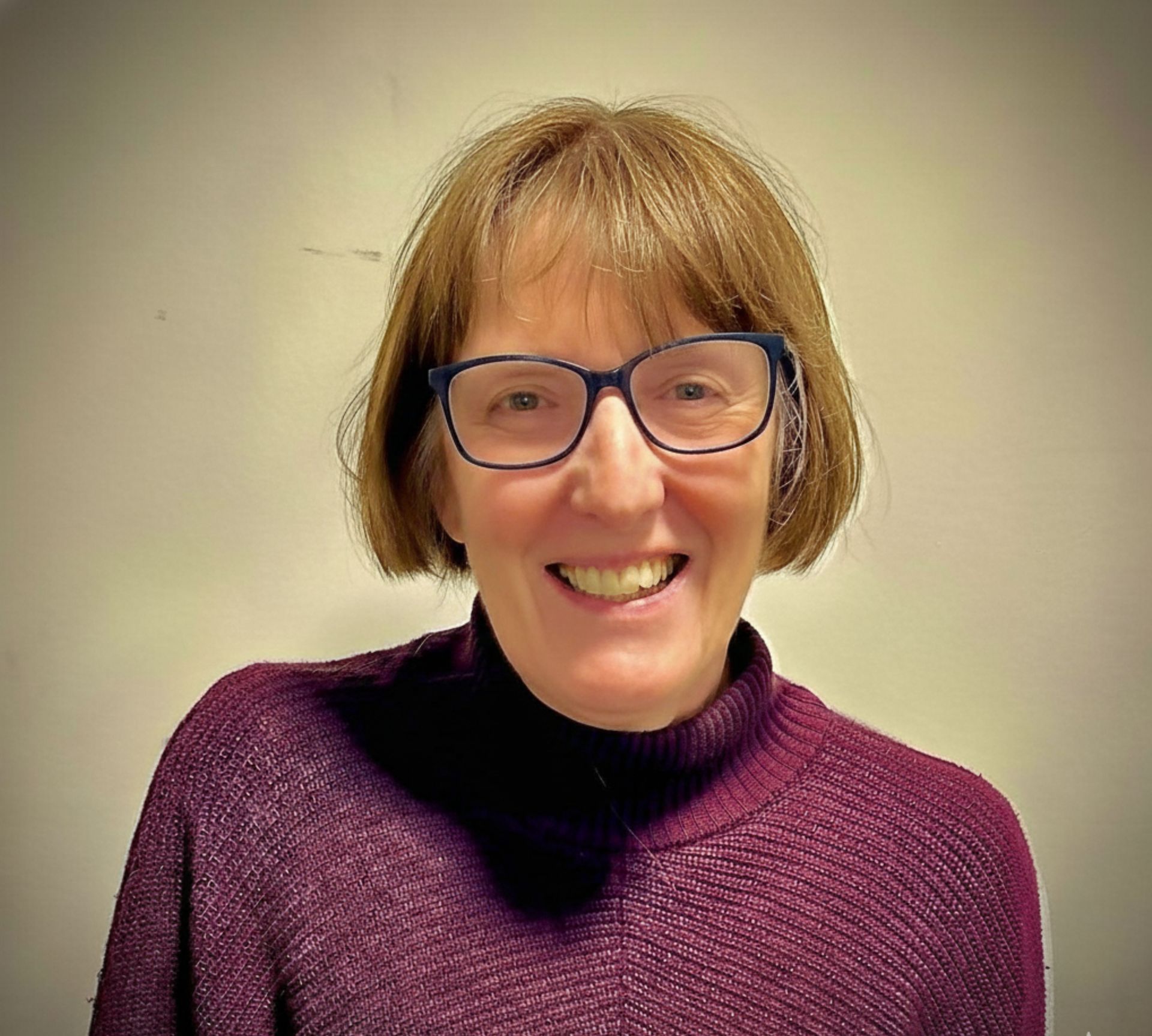 Woman with short gray hair and glasses smiles, wearing a patterned brown top.