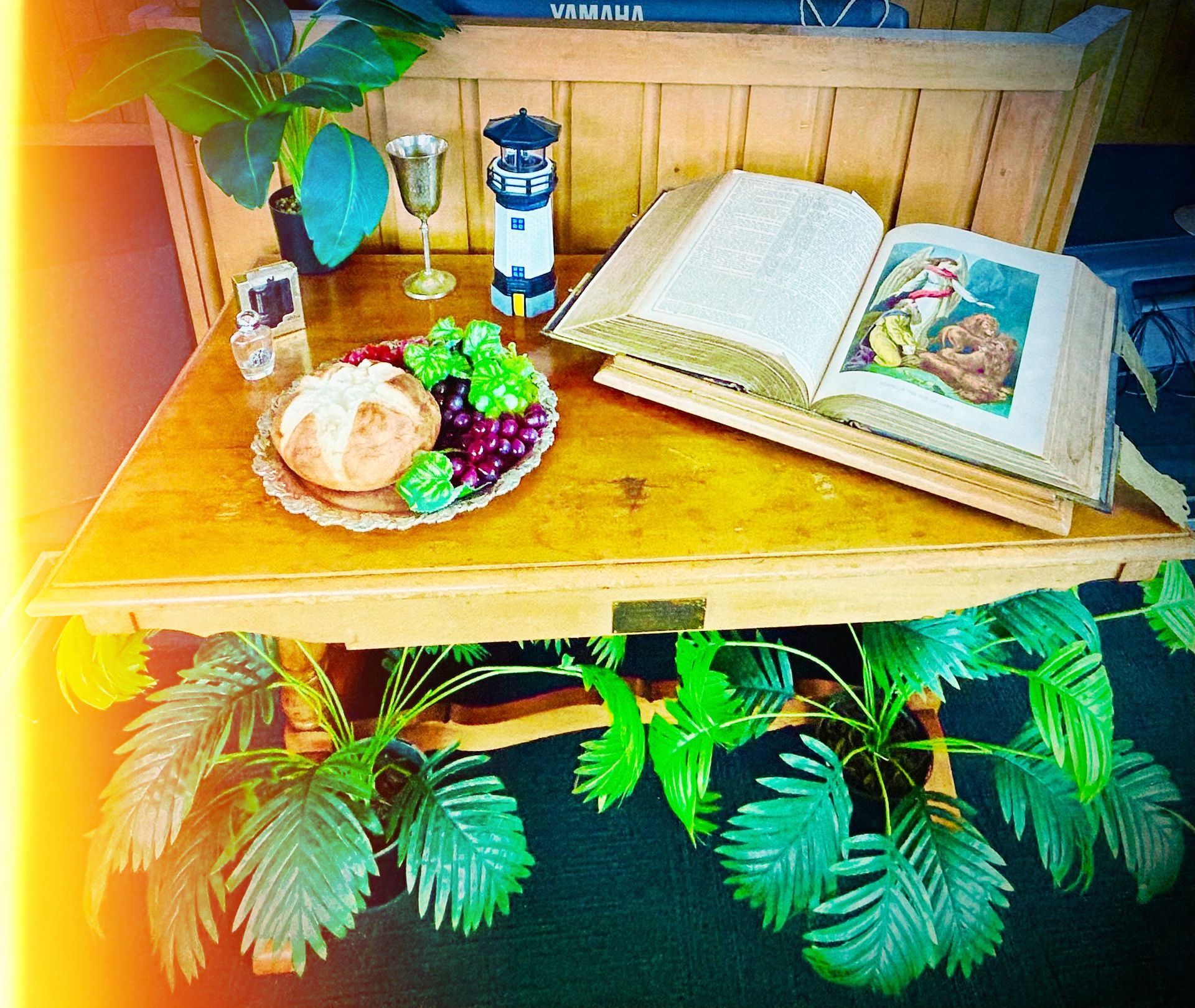 Wooden table with bread, grapes, book, and a lighthouse figurine; plants below and behind.
