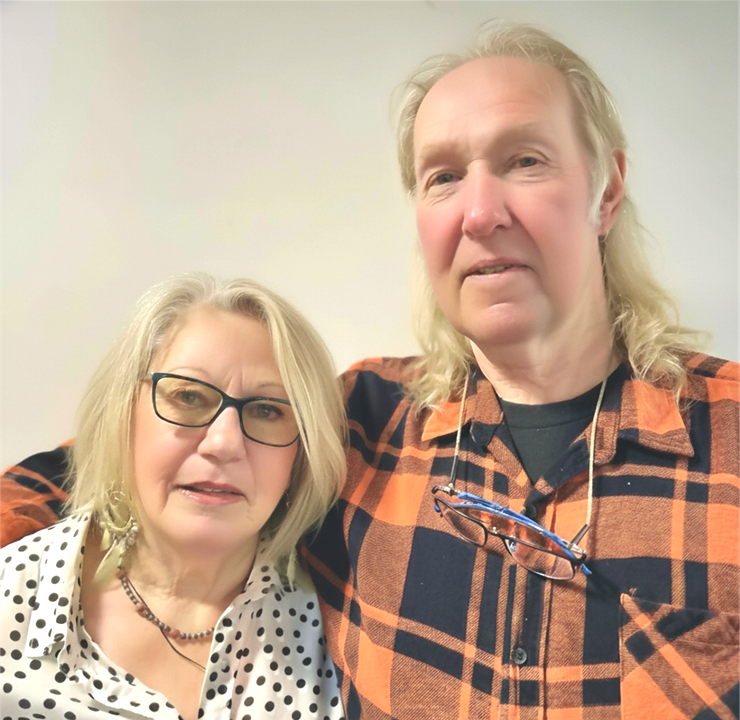 Older couple smiling. Man with beard, gray sweatshirt. Woman with glasses, patterned blouse. Beige background.