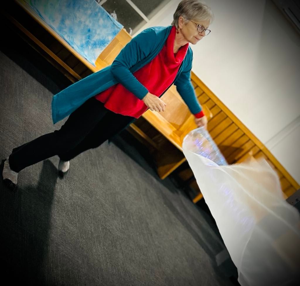 Woman in red shirt and teal sweater, holds a sheer fabric in a room with wooden benches, and gray floor.