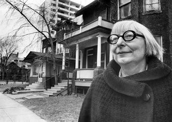 A woman wearing glasses is standing in front of a house.