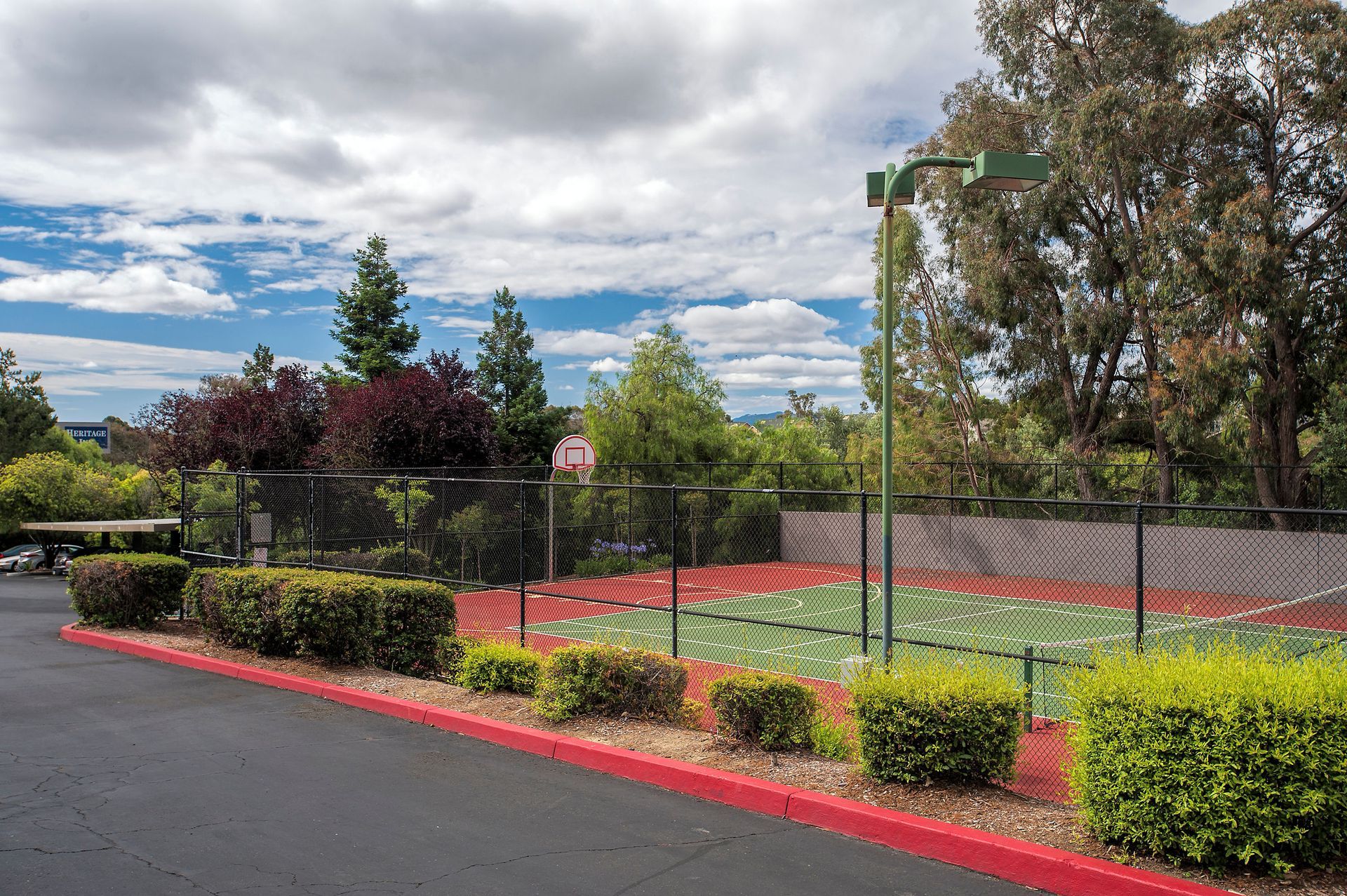 Outdoor fenced basketball court with a hoop, surrounded by shrubs and trees.