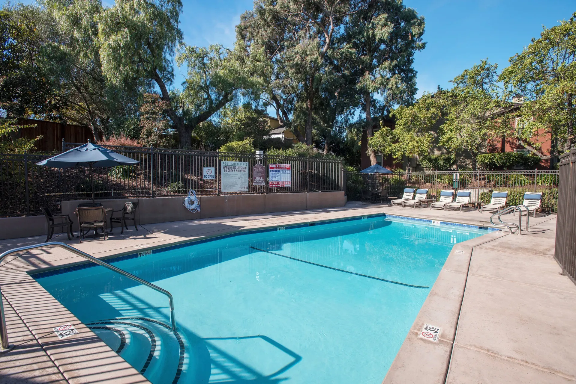 Outdoor apartment community pool with lounge chairs, tables, umbrellas, and surrounding trees.