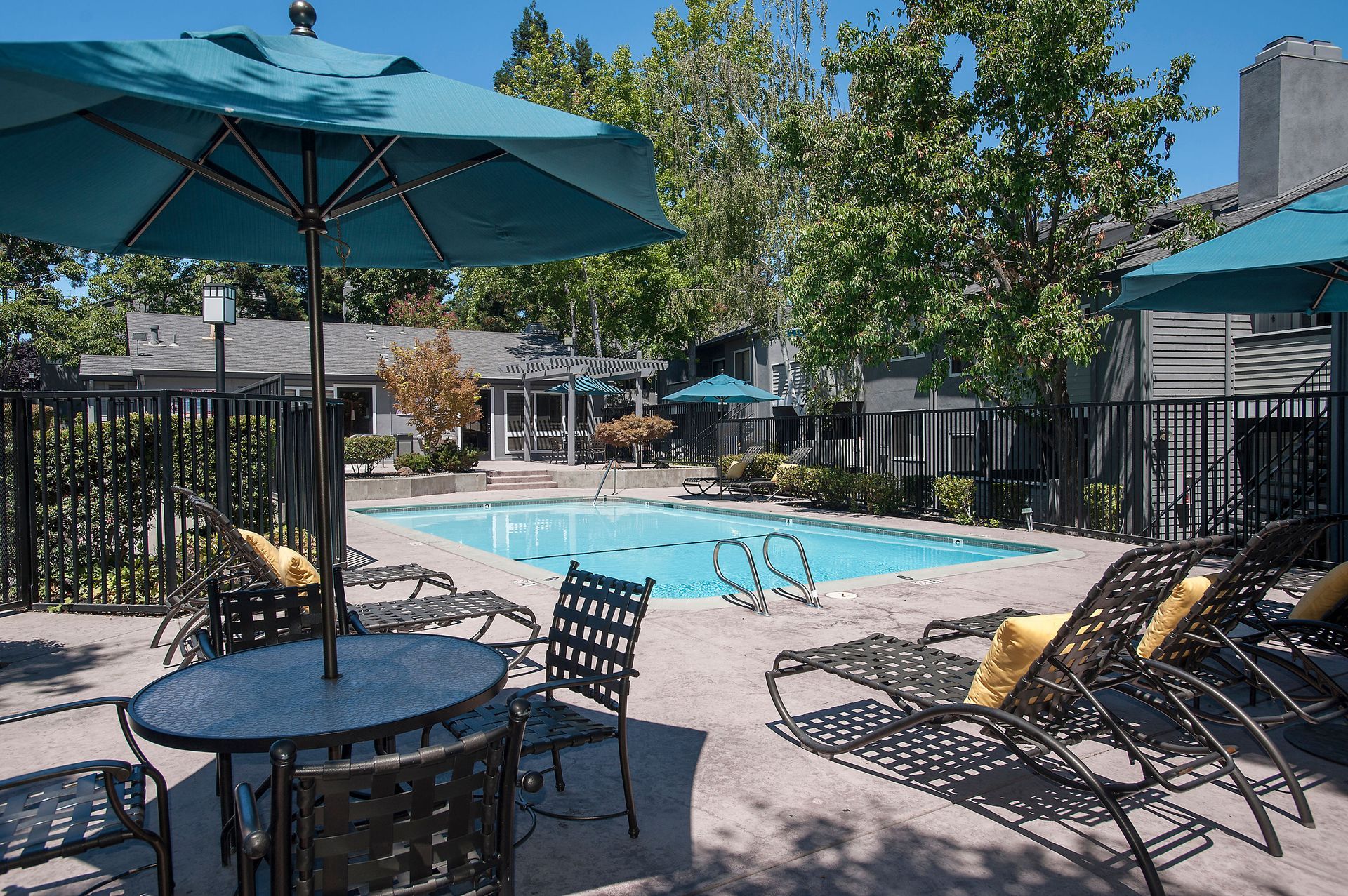 Outdoor apartment community pool with lounge chairs, teal umbrellas, and fencing.