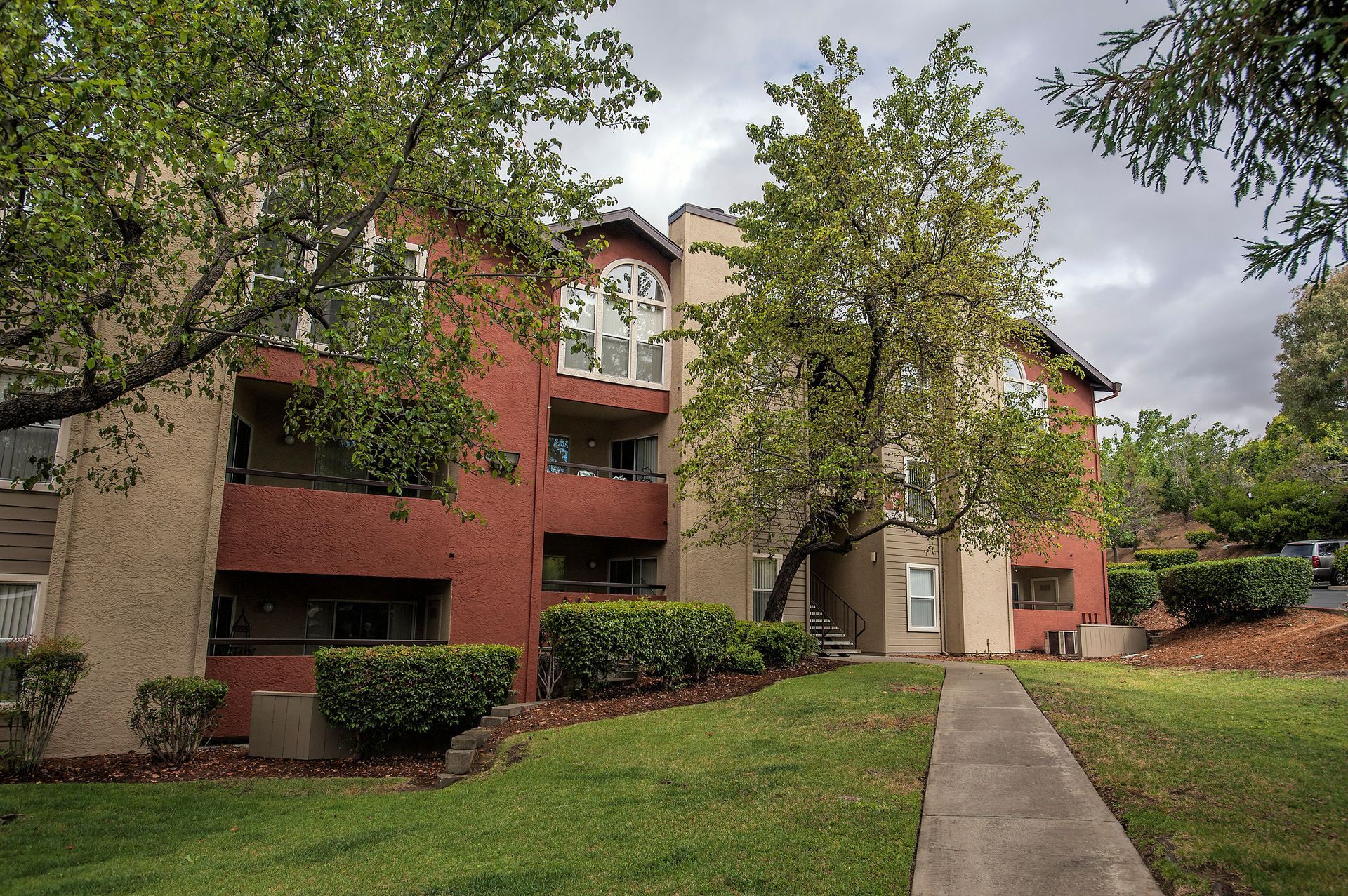 Exterior view of a multi-family building with balconies, trees, and a walkway.