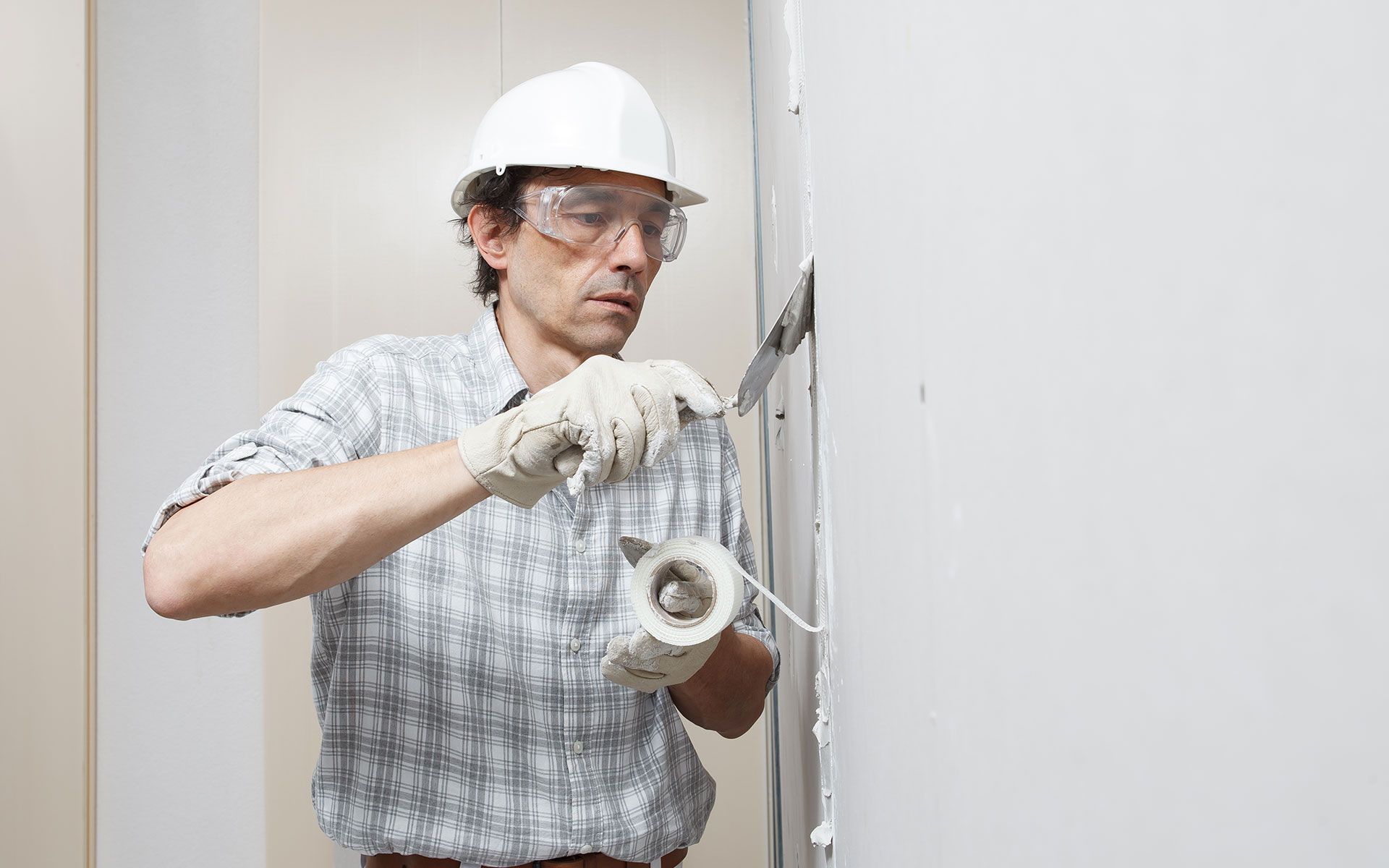 Construction worker wearing a hard hat and safety glasses, applying tape to a wall.