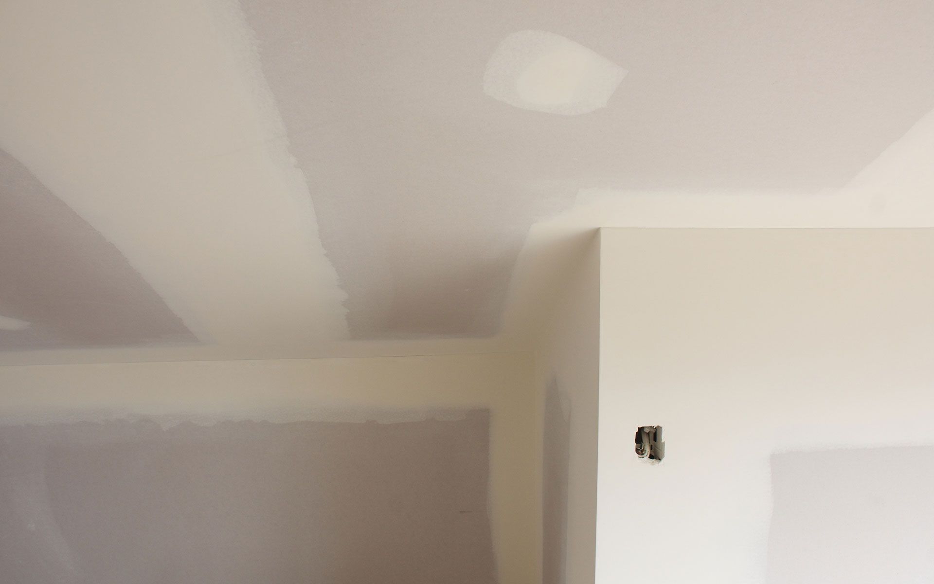 Drywall ceiling and walls in a room; beige and off-white colors, unfinished. Small patch and electrical box visible.