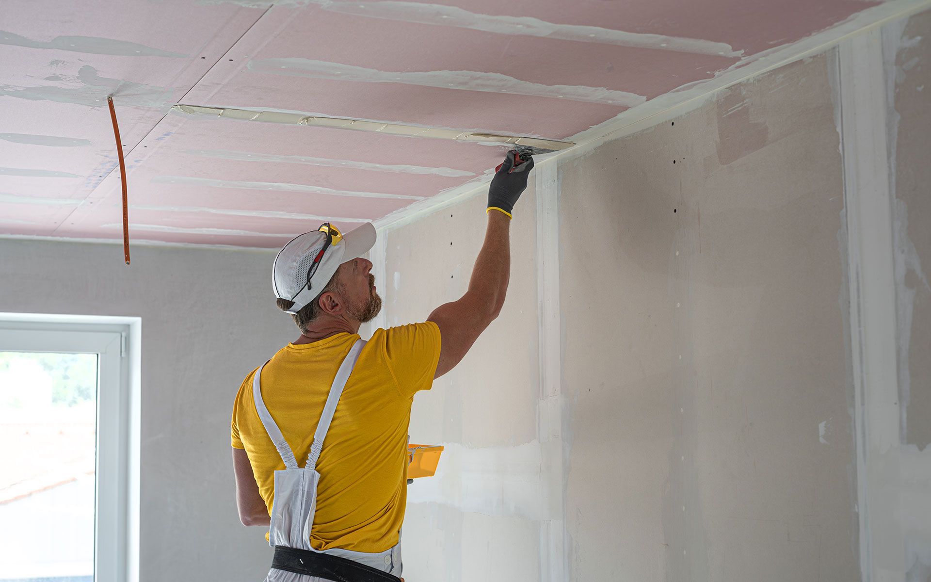 Man in yellow shirt and hard hat, applying joint compound to drywall seams on ceiling and wall. Indoors, construction site.
