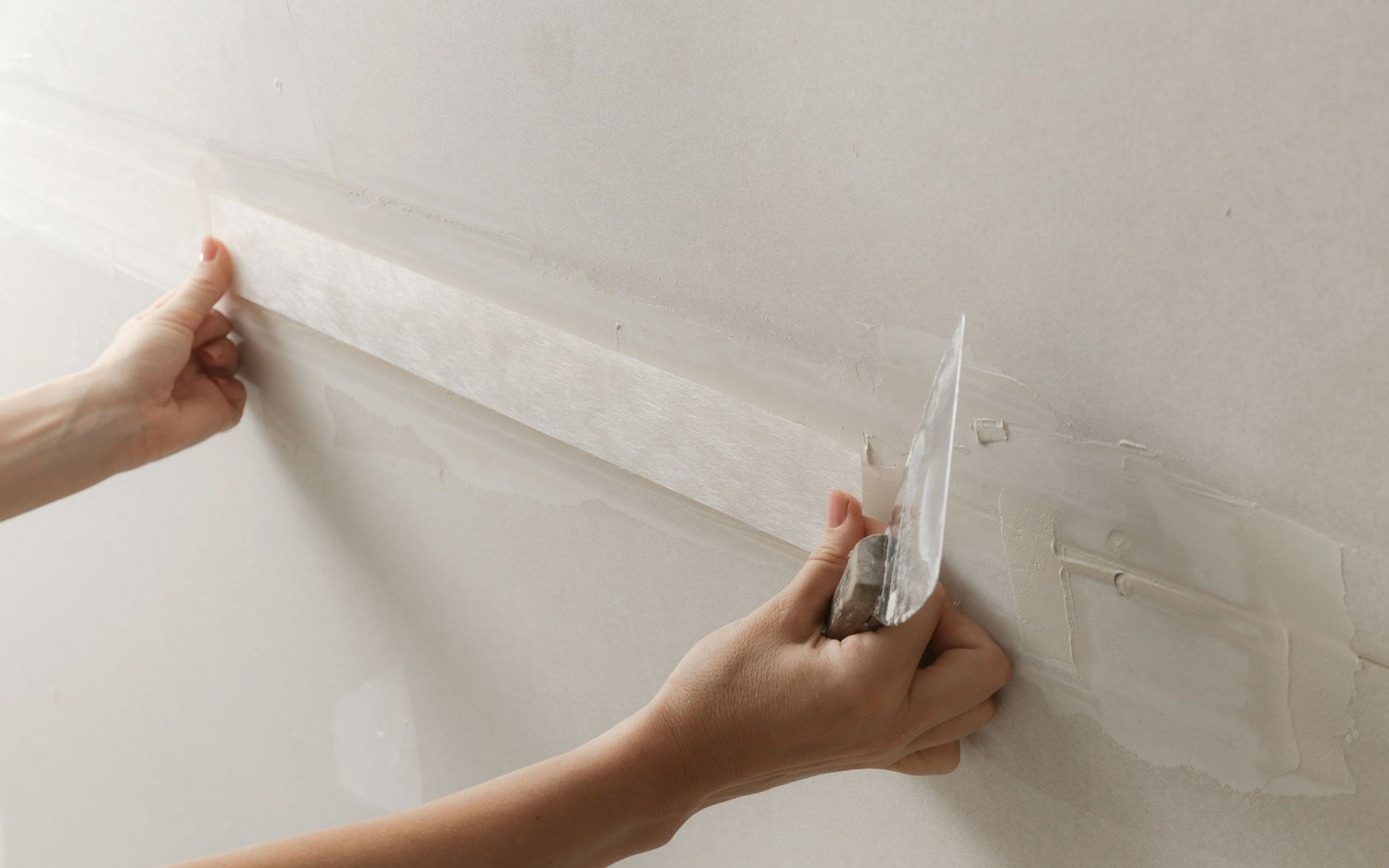 Person applying drywall tape to a wall with a putty knife, indoors. Hands are visible.