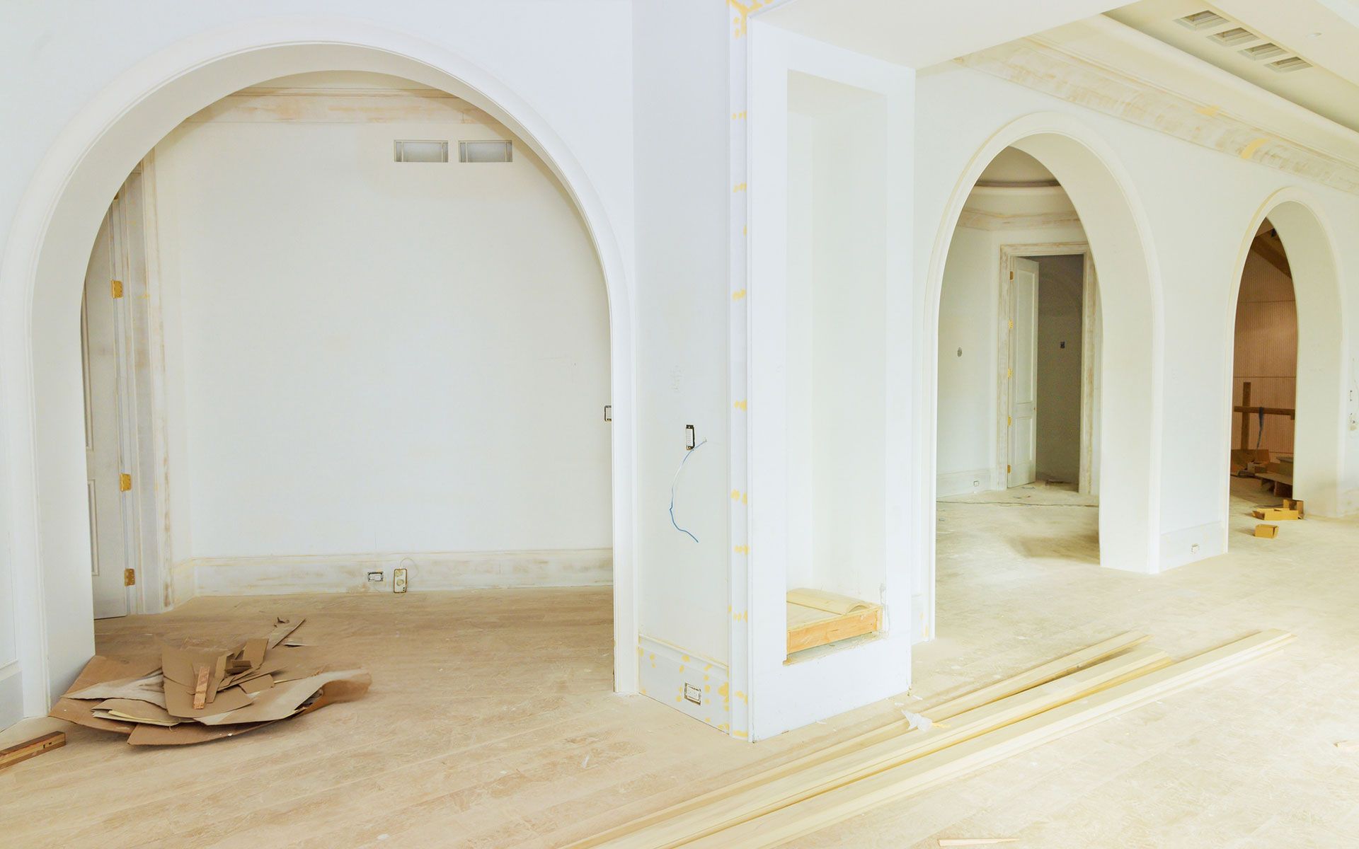Interior view of a room with arched doorways and white walls during renovation. Wood flooring and construction materials are visible.
