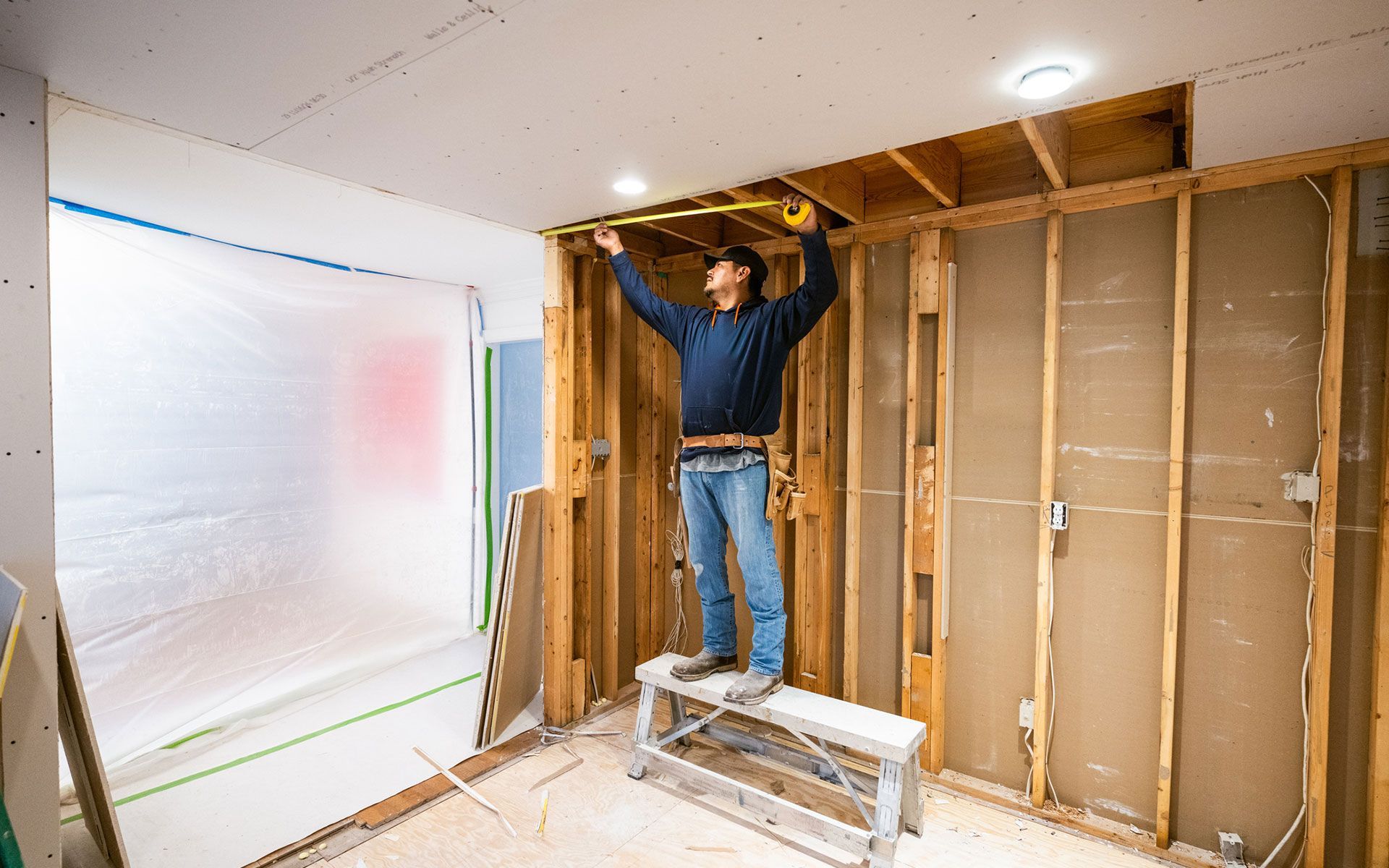 A person on a step stool measures the ceiling during a room renovation, surrounded by exposed framing and unfinished walls.