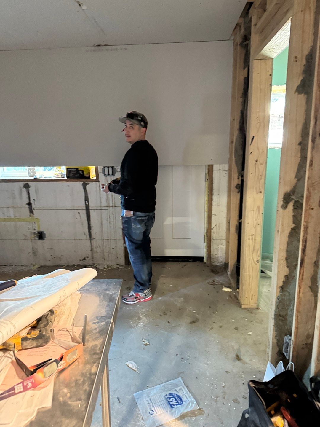 A person in a construction zone, wearing a cap and jeans, stands near a wall with partially installed paneling and exposed framing.
