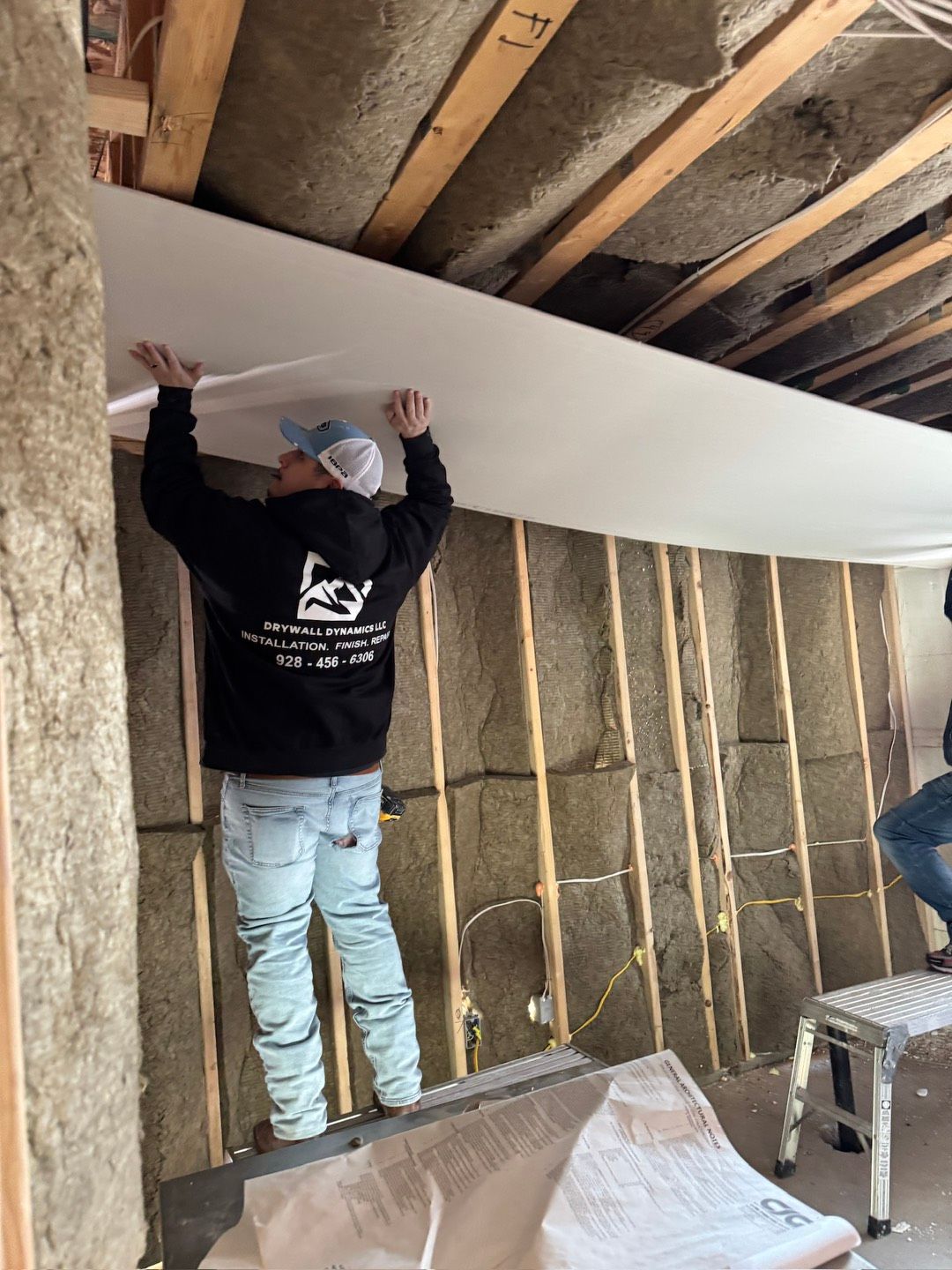 A person in a black hoodie installs ceiling material in a room under construction. Walls have insulation, wood beams are visible.