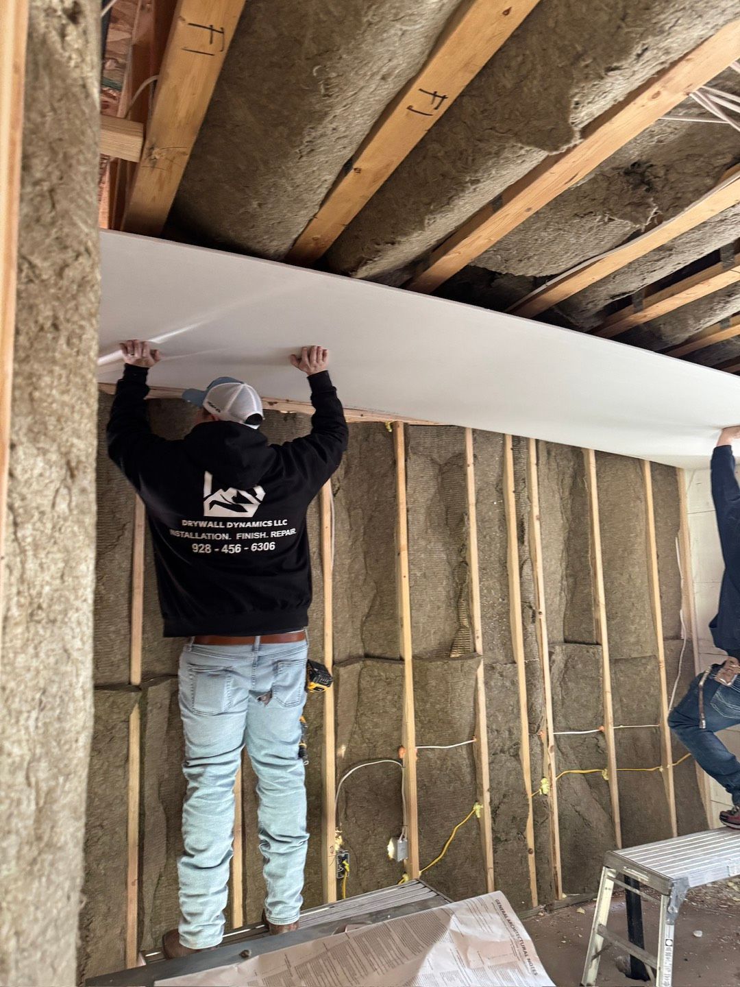 Two construction workers installing drywall on a ceiling. The setting is a room with exposed studs and insulation.