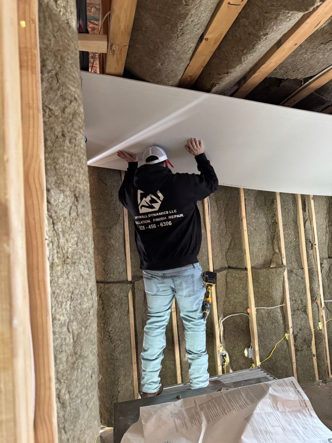 A person wearing a black hoodie and jeans installs drywall on a ceiling. The room is under construction with insulation visible.