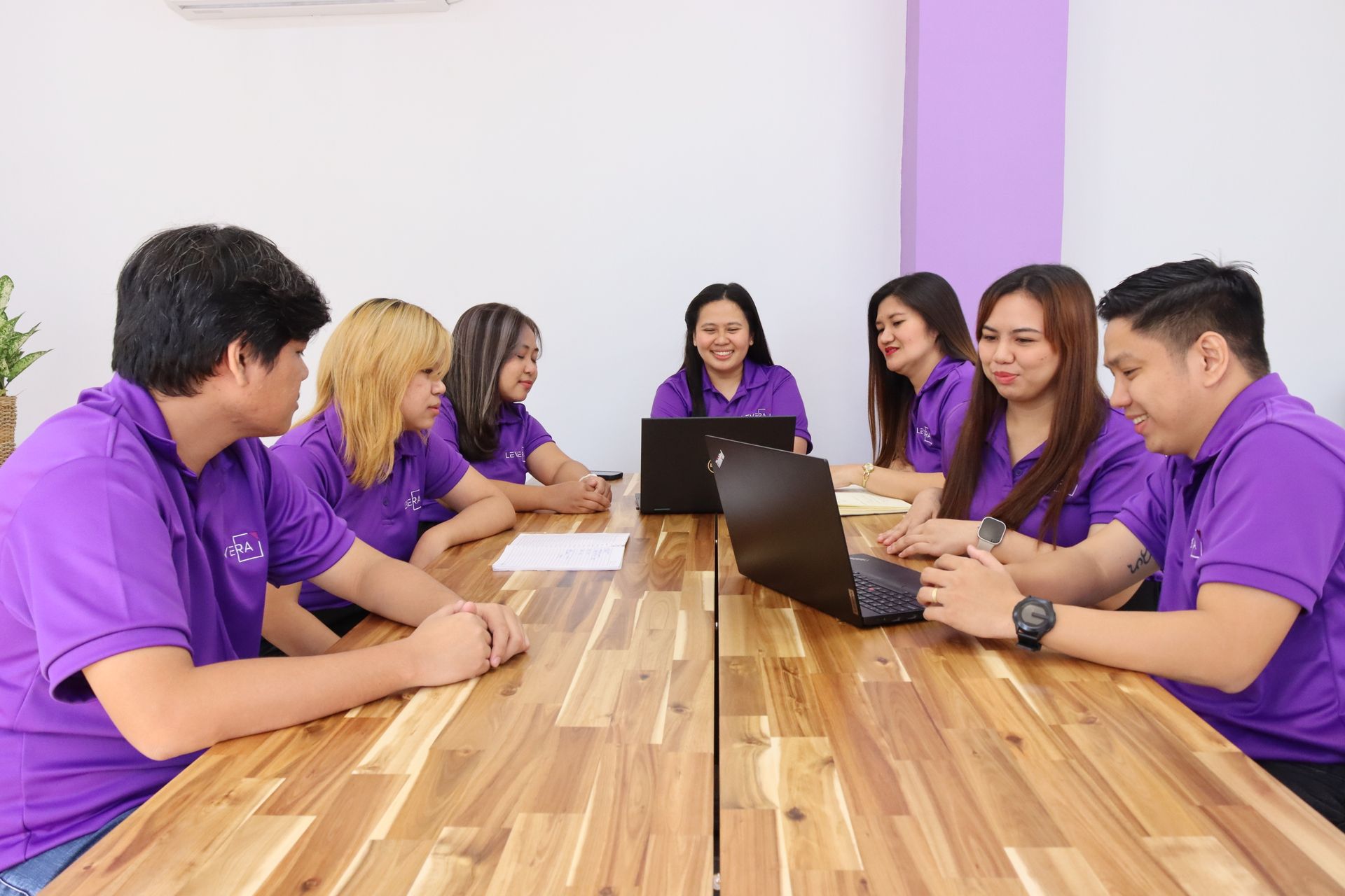A group of people are sitting around a wooden table with laptops.