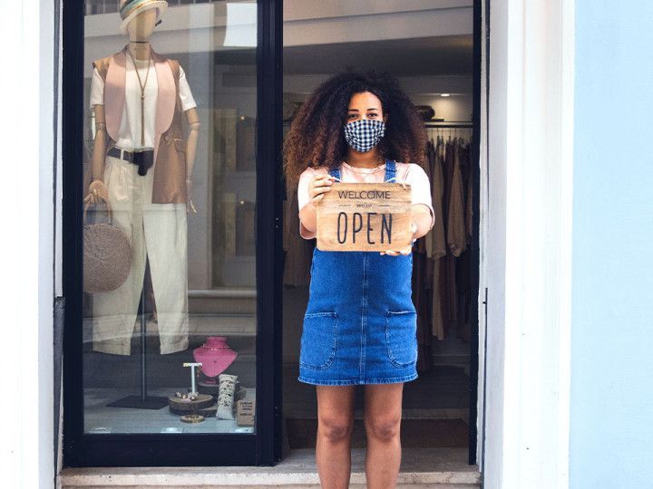 A woman wearing a mask is holding an open sign in front of a store.