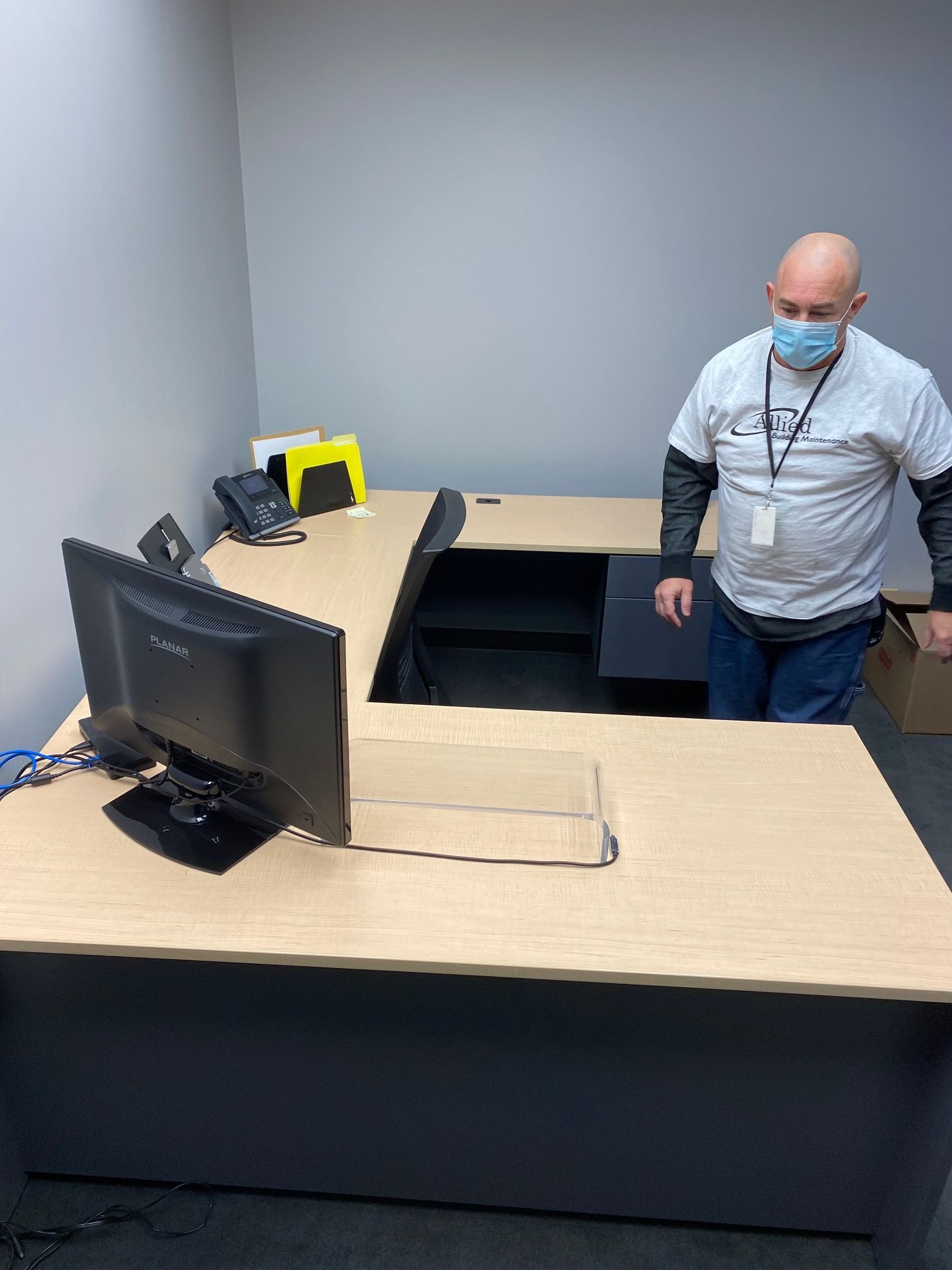 A man wearing a mask is standing in front of a desk with a computer on it.