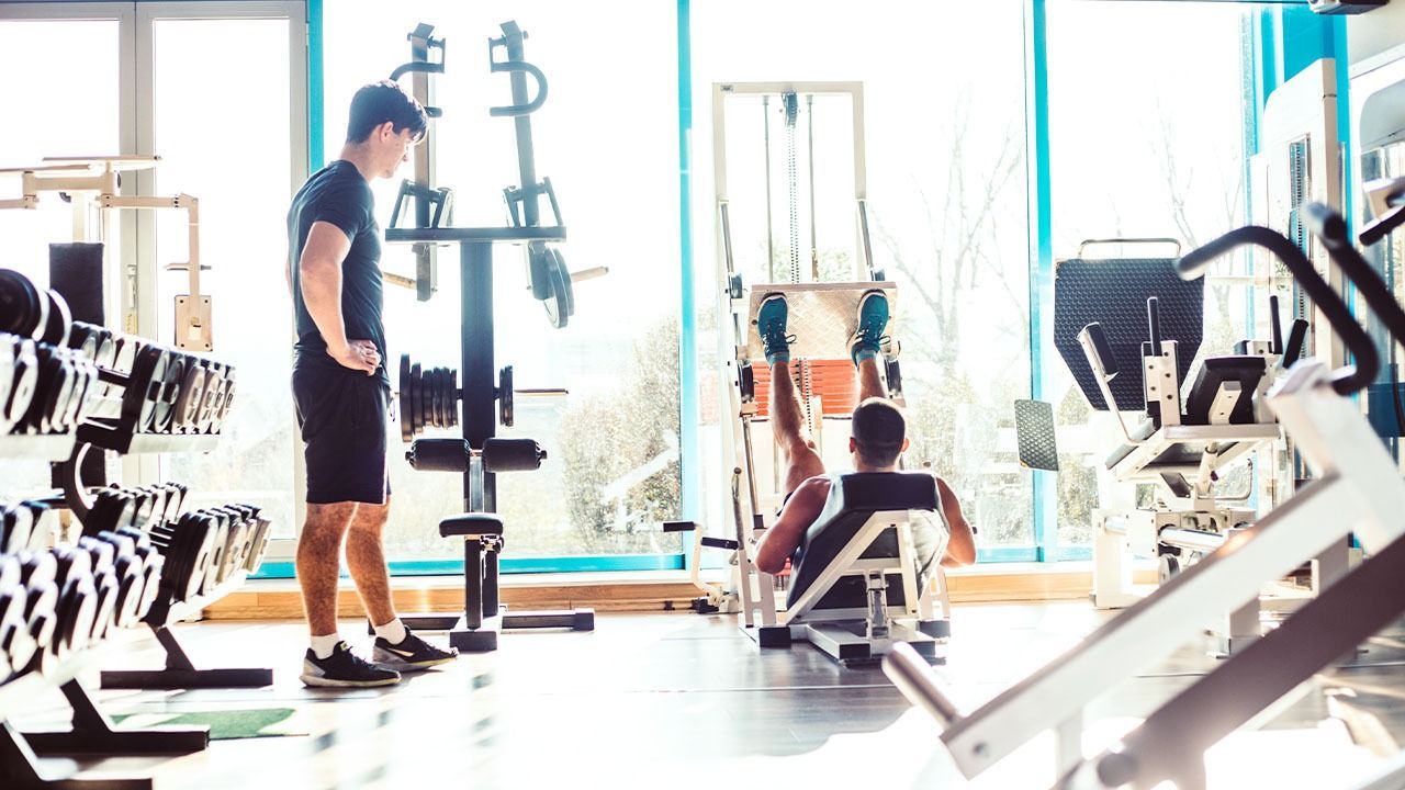A man is standing in a gym next to a man sitting on a machine.