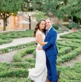 Couple embracing in a garden maze, dressed in white and blue, smiling at the camera.