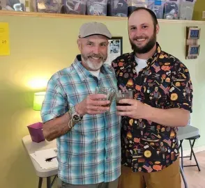 Two smiling people holding drinks indoors at a casual party, with a lamp and table behind them