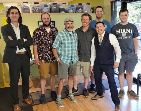 Six people posing indoors in front of shelves, wearing casual and business-casual clothes