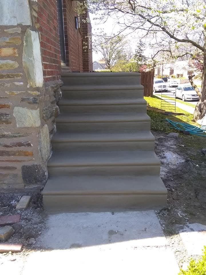 Concrete stairs beside a brick house leading up from a sidewalk, with a street and parked cars nearby