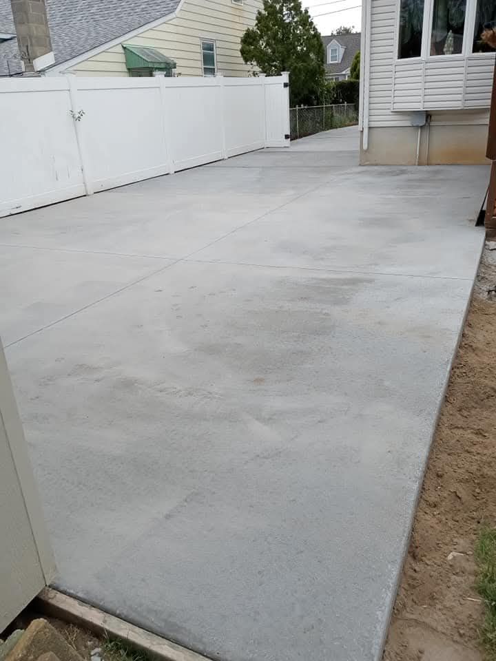 Concrete patio beside a house with a white fence and narrow dirt strip along the edge